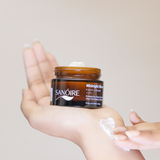 Hand holding Sanoire Midnight Bloom face cream jar with cream scooped on fingers