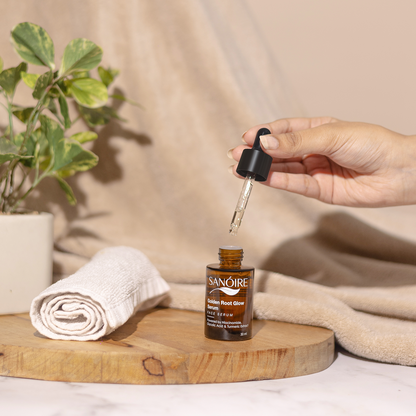 Hand holding dropper above Sanoire Golden Root Glow Serum bottle on wooden tray, with towel and plant.
