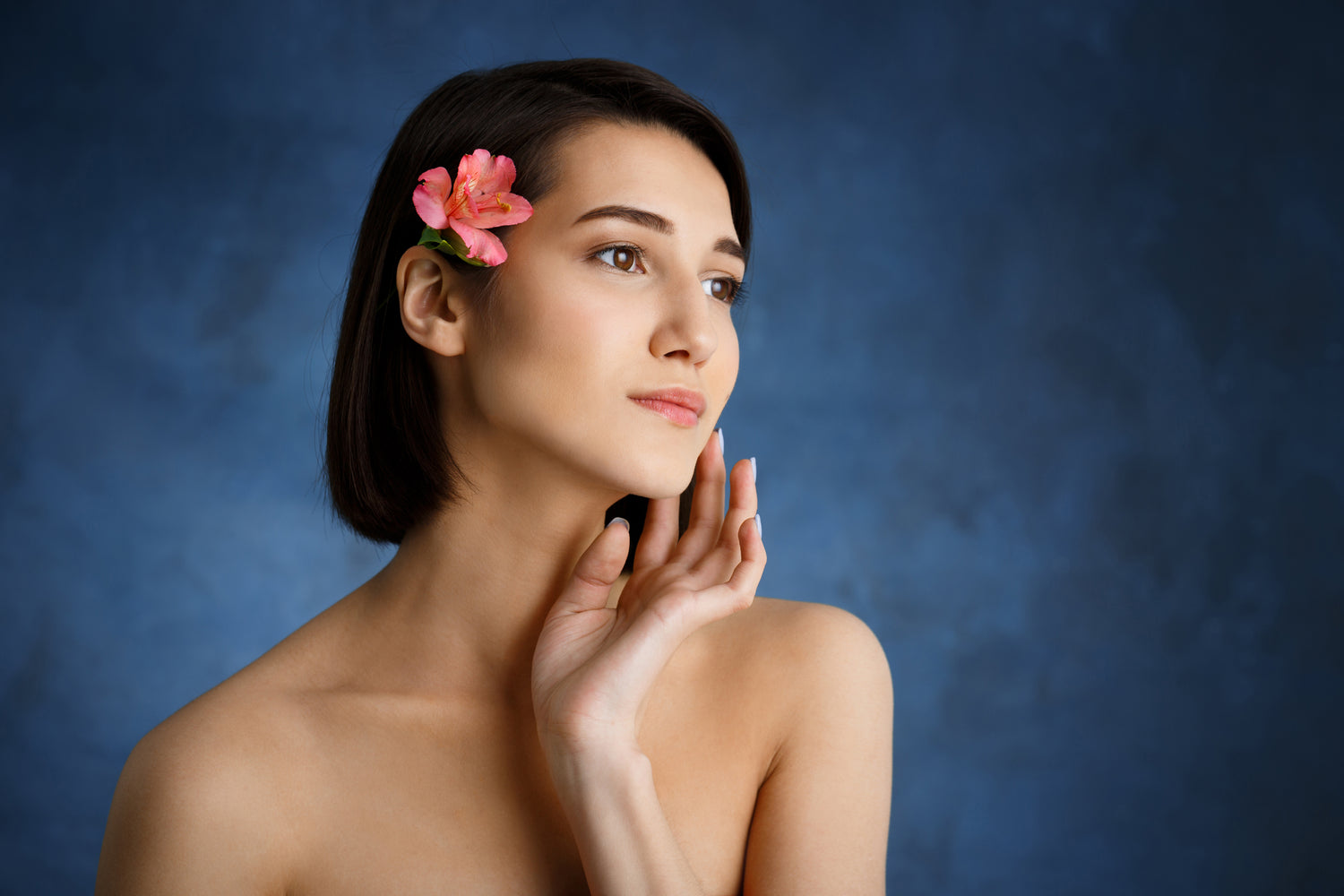 Young woman with radiant bare skin and pink flower in hair against a blue background